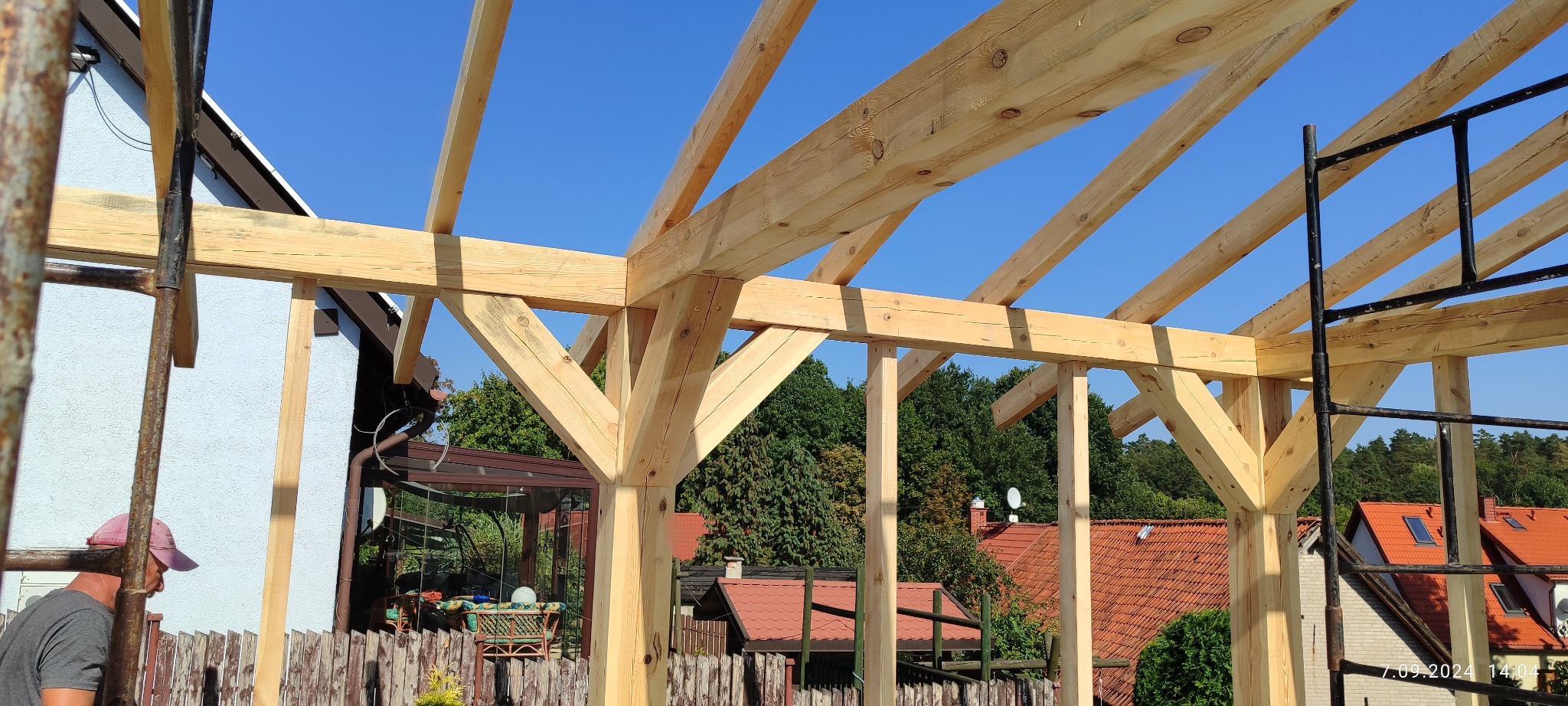 Drewniana konstrukcja altany w trakcie budowy, widoczne elementy nośne i złączenia ciesielskie na tle błękitnego nieba i zieleni drzew. W tle zabudowa jednorodzinna.