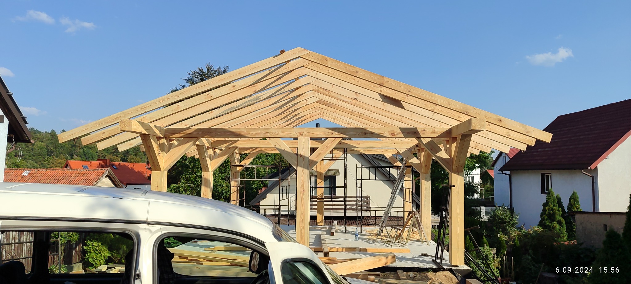 Konstrukcja drewniana altany w trakcie budowy, widoczne elementy więźby dachowej i słupy nośne. W tle budynki mieszkalne i zieleń.