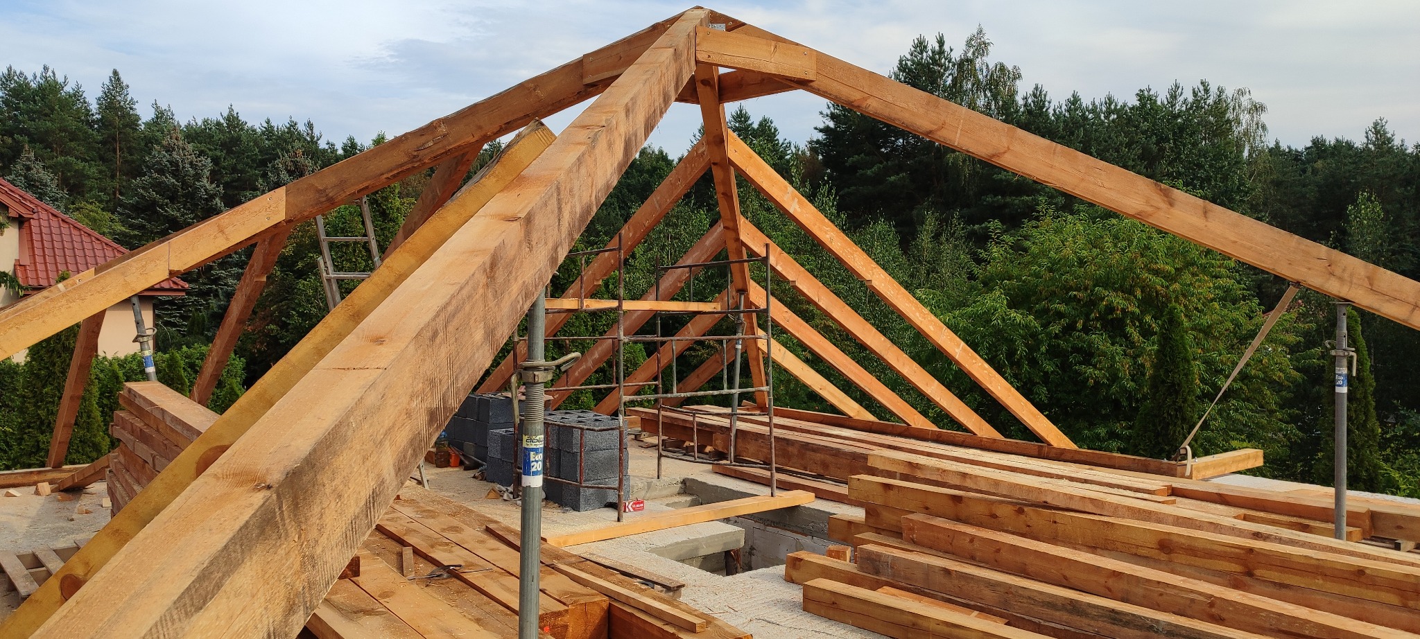 Konstrukcja drewniana więźby dachowej w trakcie montażu, widok z góry, na tle zieleni. Elementy wsparte na podporach. Częściowo widoczny budynek z czerwoną dachówką.