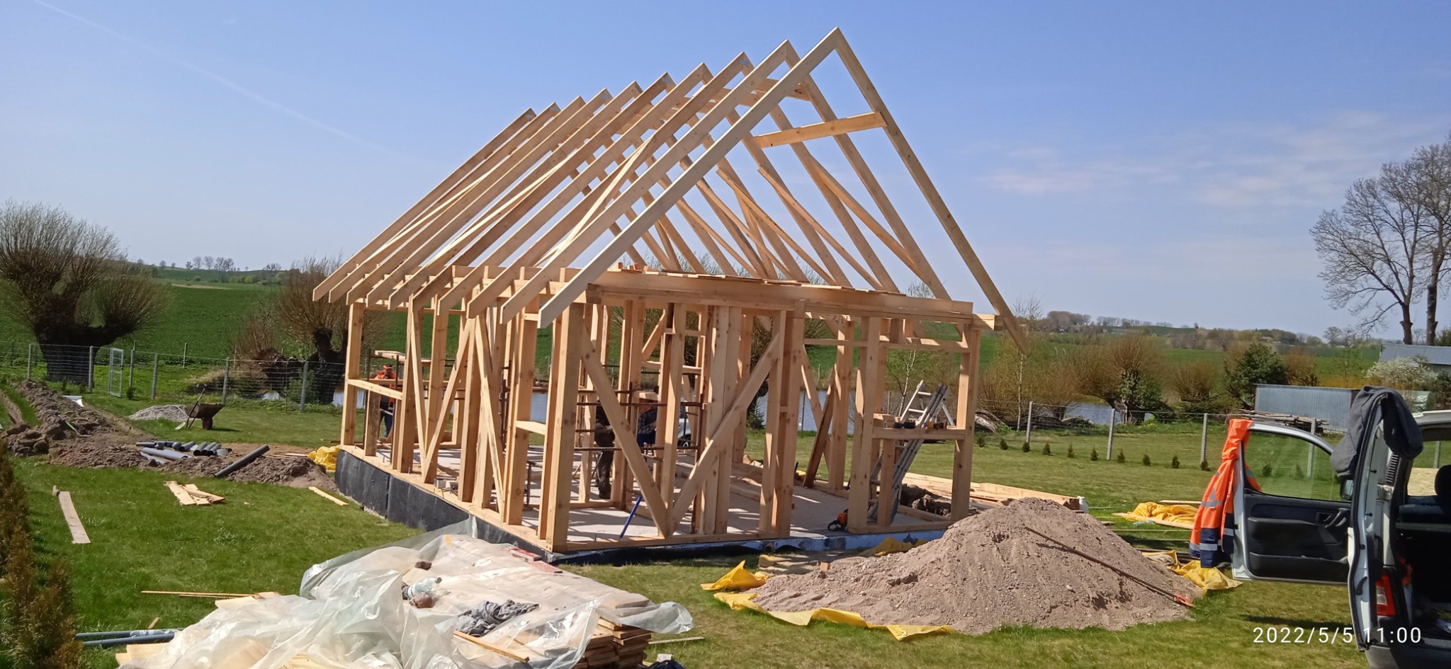 Konstrukcja szkieletowa domu w trakcie budowy, widoczna więźba dachowa na tle zielonego krajobrazu i błękitnego nieba. Etap montażu belek.