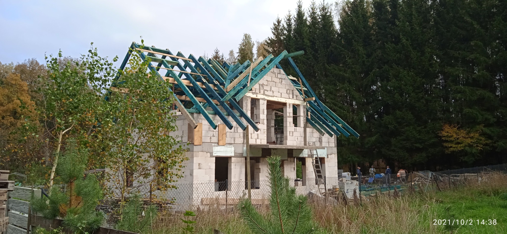 Surowy dom z bloczków z niebieską więźbą dachową na tle lasu iglastego, widoczny przez ogrodzenie. Budowa w toku, jesienna sceneria.