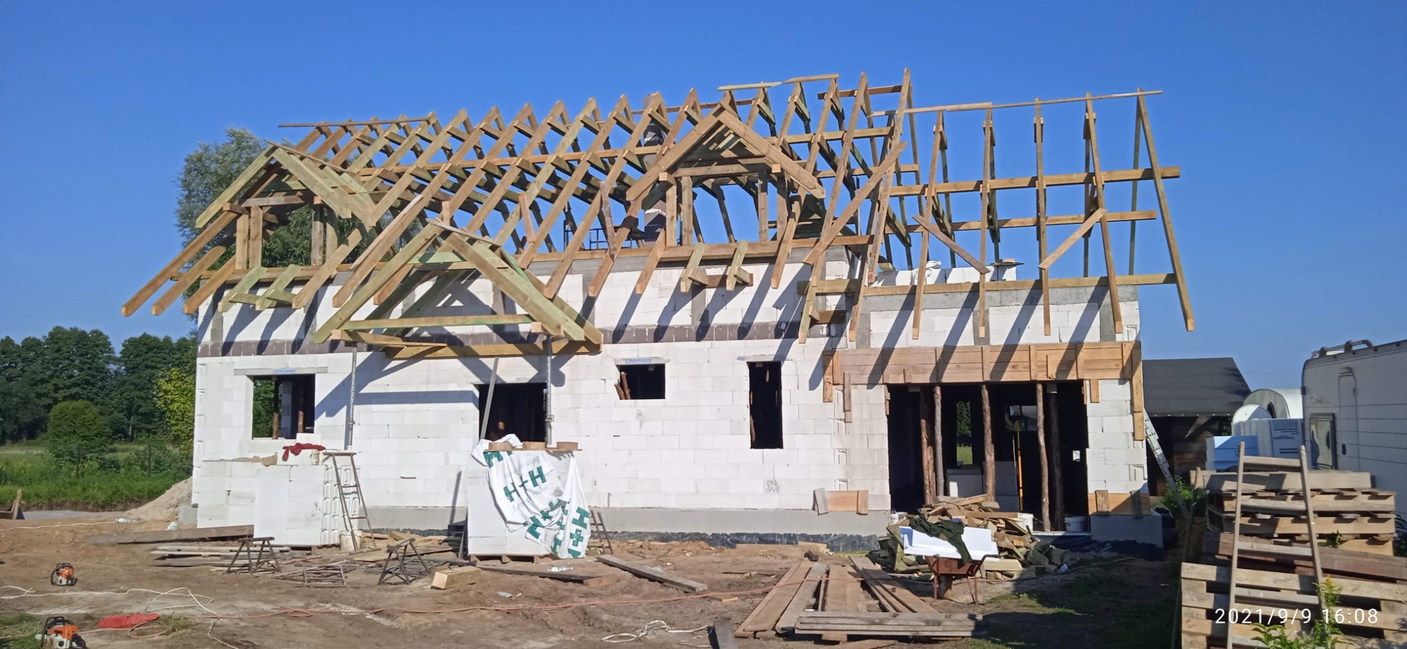 Budowa domu z białego pustaka: wzniesiona więźba dachowa z drewnianych belek na tle błękitnego nieba. Widoczne narzędzia i materiały budowlane na placu budowy.