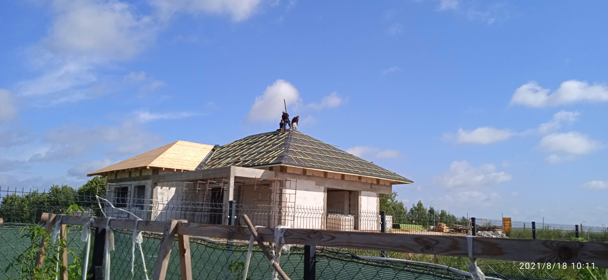 Montaż łat na dachu domu jednorodzinnego w trakcie budowy. Na dachu pracują dwie osoby. Część dachu pokryta deskami, część folią i łatami. Błękitne niebo z chmurami w tle.