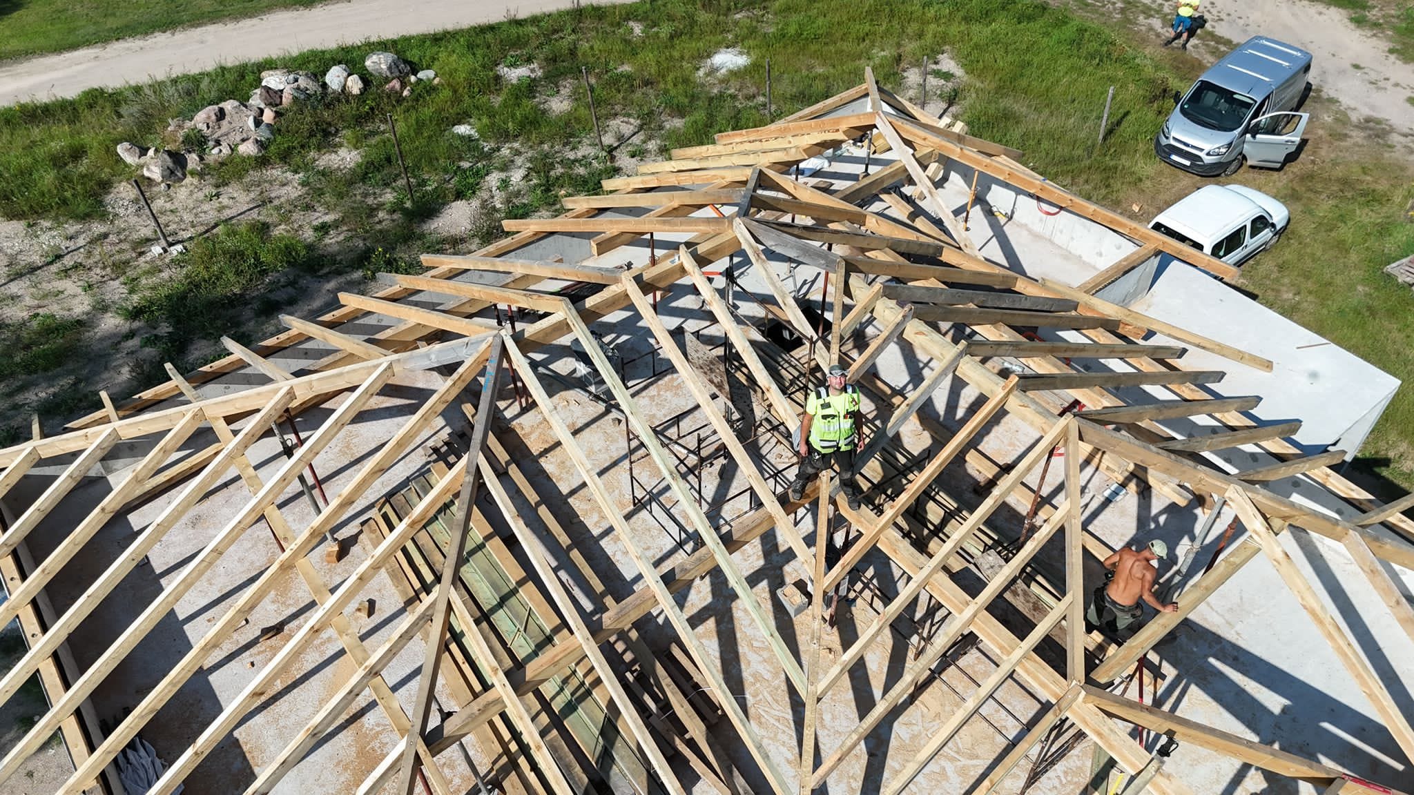 Montaż drewnianej więźby dachowej z perspektywy drona. Dwóch pracowników w trakcie pracy. Uwagę przykuwa precyzja wykonania i jasne drewno konstrukcyjne.