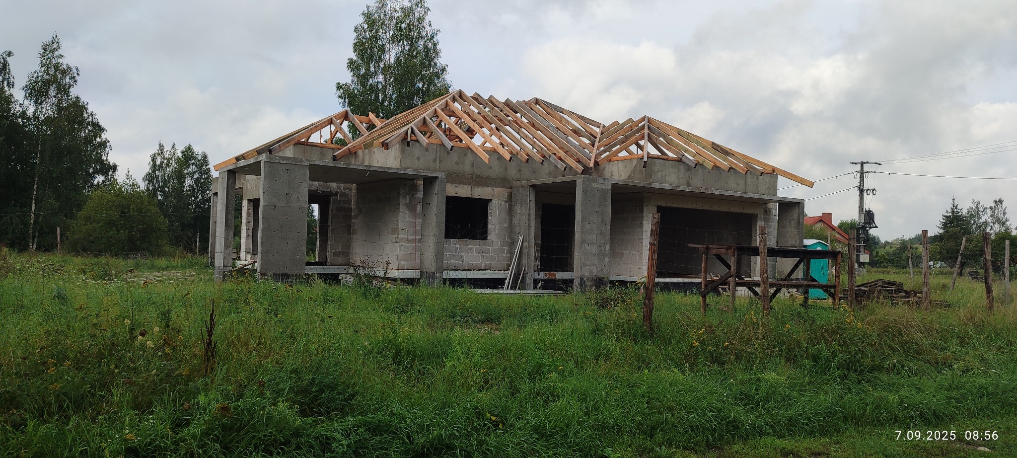 Budowa domu w stanie surowym otwartym: ściany z betonu komórkowego i drewniana konstrukcja dachu. Widok na zarośniętą działkę i pochmurne niebo.