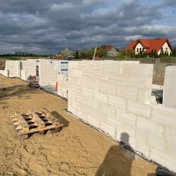 KWARCIAK BUD - Wznoszenie ścian z bloczków Solbet na placu budowy domu jednorodzinnego, widoczny fragment fundamentów, paleta z materiałem i cień osoby fotografującej na piaszczystym podłożu.