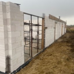 KWARCIAK BUD - Wznoszenie ścian domu z białych bloczków, widoczne rusztowania i zbrojenia, teren wokół w stanie surowym, przygotowany pod dalsze prace.