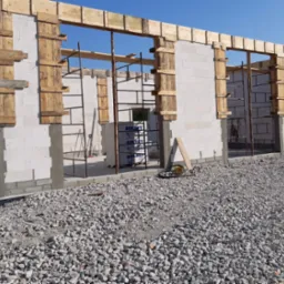 Budowa domu z białych bloczków: ściany w trakcie wznoszenia, widoczne drewniane szalunki okienne, stalowe rusztowania i paleta bloczków Solbet w tle.