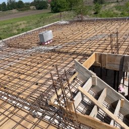 KWARCIAK BUD - Zbrojenie stropu z drewnianym szalunkiem i widokiem na zielone pola w tle.