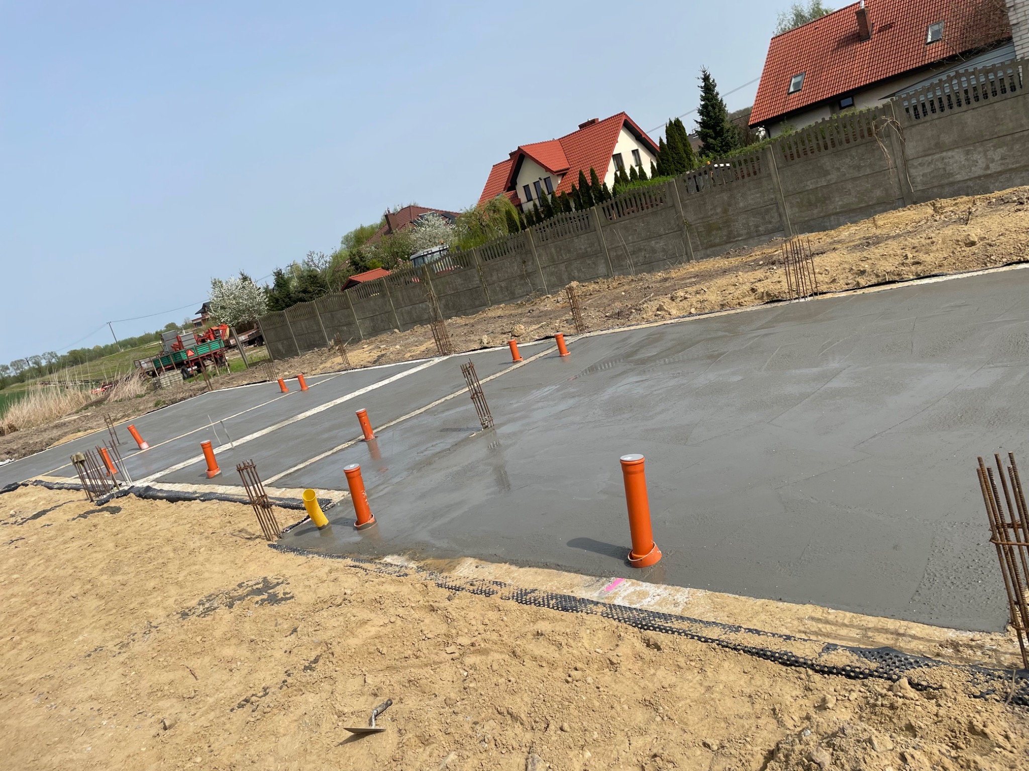 Świeżo wylana, gładka płyta fundamentowa z wystającymi prętami zbrojeniowymi i pomarańczowymi rurami pionowymi, widok na teren budowy z oddali, w tle ogrodzenie i domy.