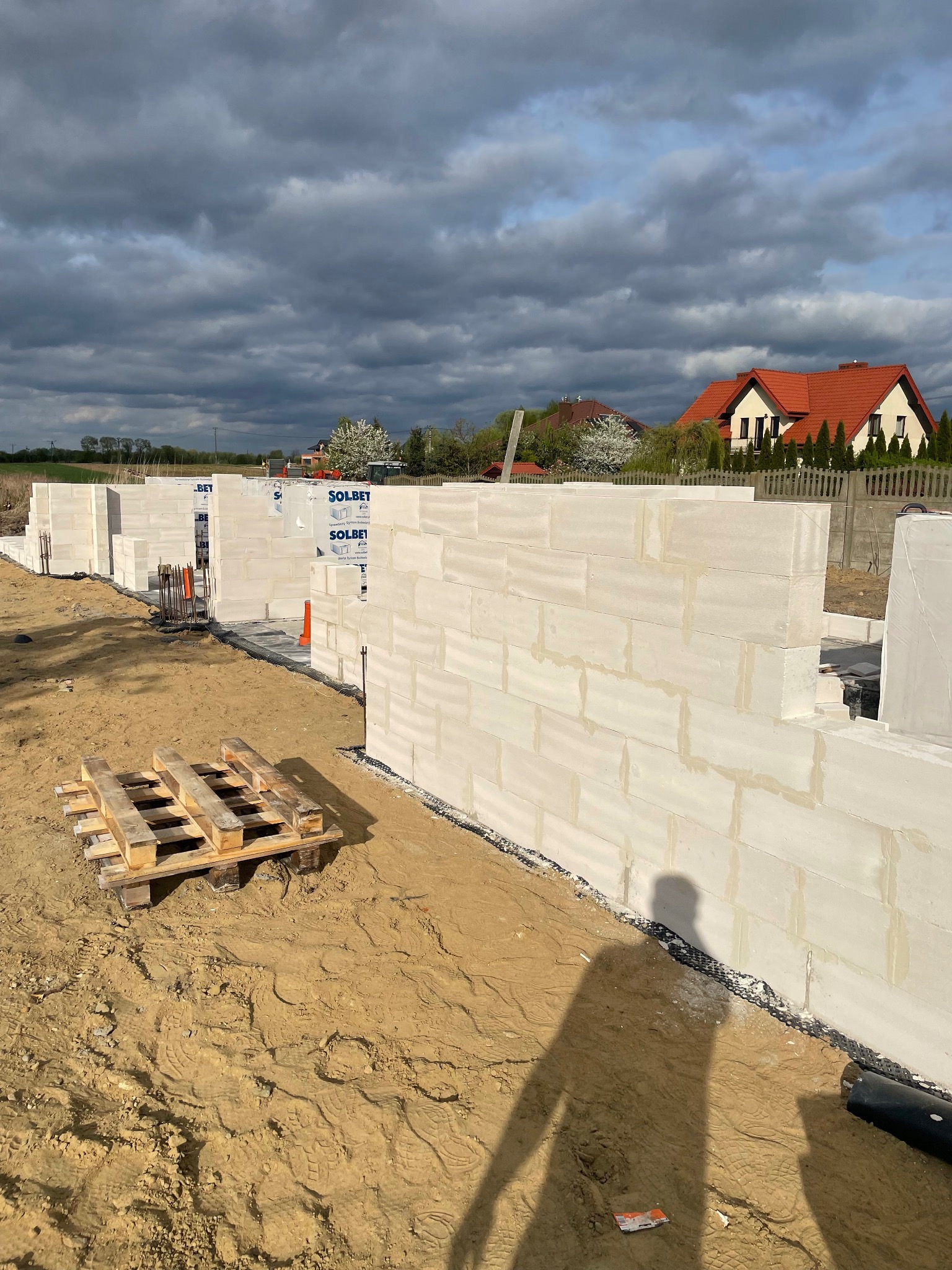 Wznoszenie ścian z bloczków Solbet na placu budowy domu jednorodzinnego, widoczny fragment fundamentów, paleta z materiałem i cień osoby fotografującej na piaszczystym podłożu.