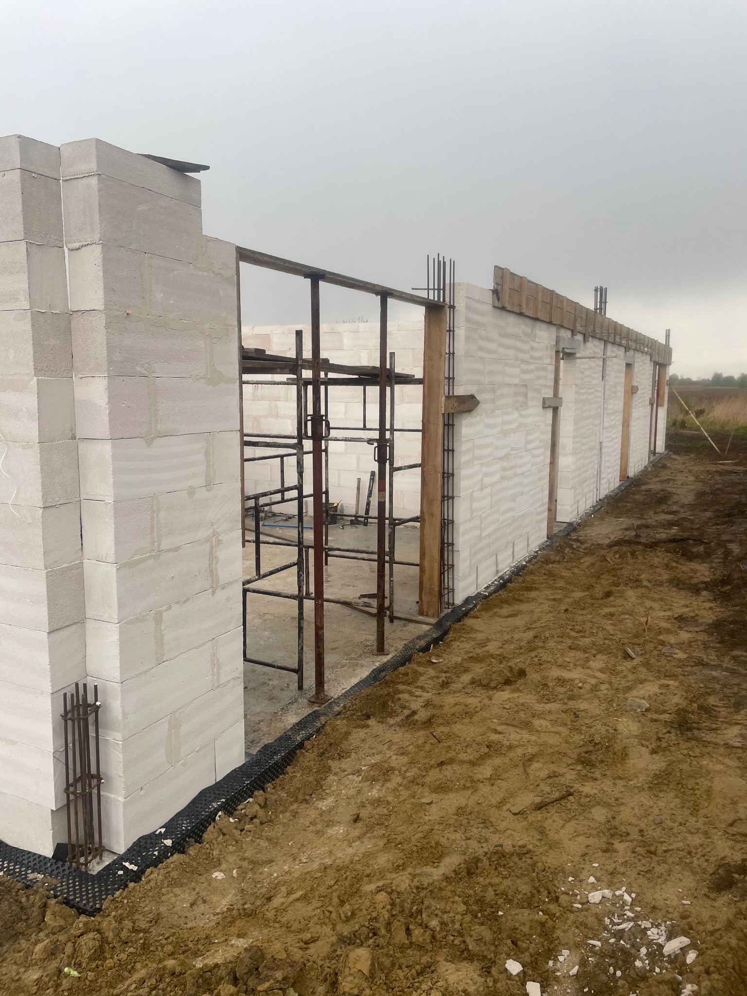 Wznoszenie ścian domu z białych bloczków, widoczne rusztowania i zbrojenia, teren wokół w stanie surowym, przygotowany pod dalsze prace.