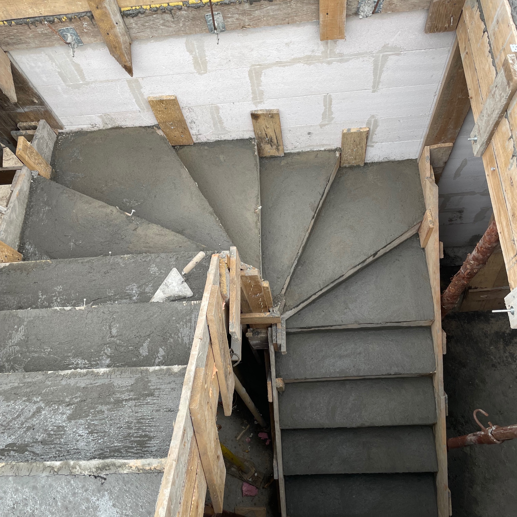 Wewnętrzne schody spiralne w trakcie wylewania betonem, z widocznym szalunkiem drewnianym i kielnią murarską na jednym ze stopni, beton jeszcze mokry.