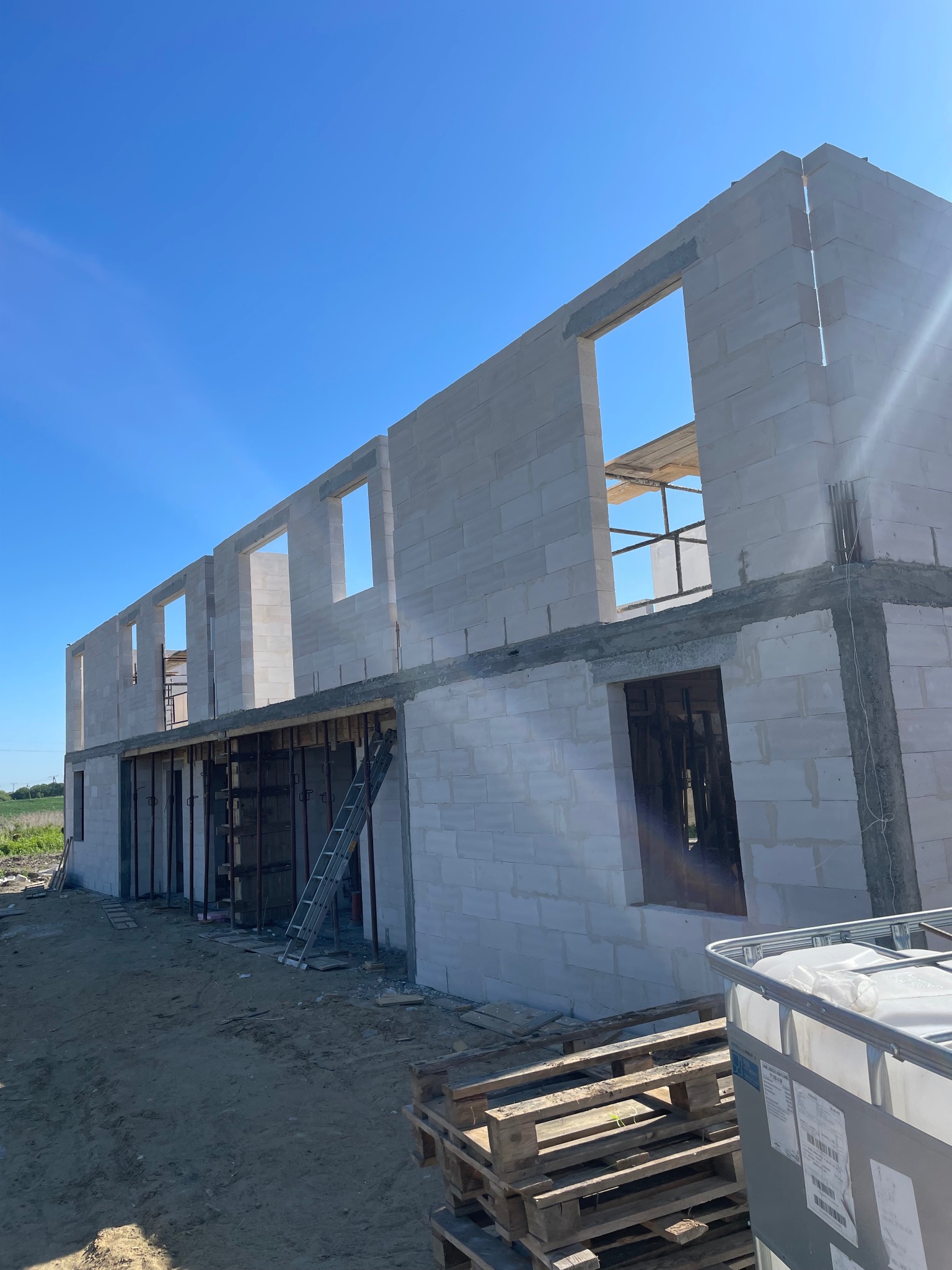 Wznoszenie dwukondygnacyjnego budynku z białych bloczków, widoczne otwory okienne, szalunki na parterze i drabina oparta o ścianę, błękitne niebo w tle.