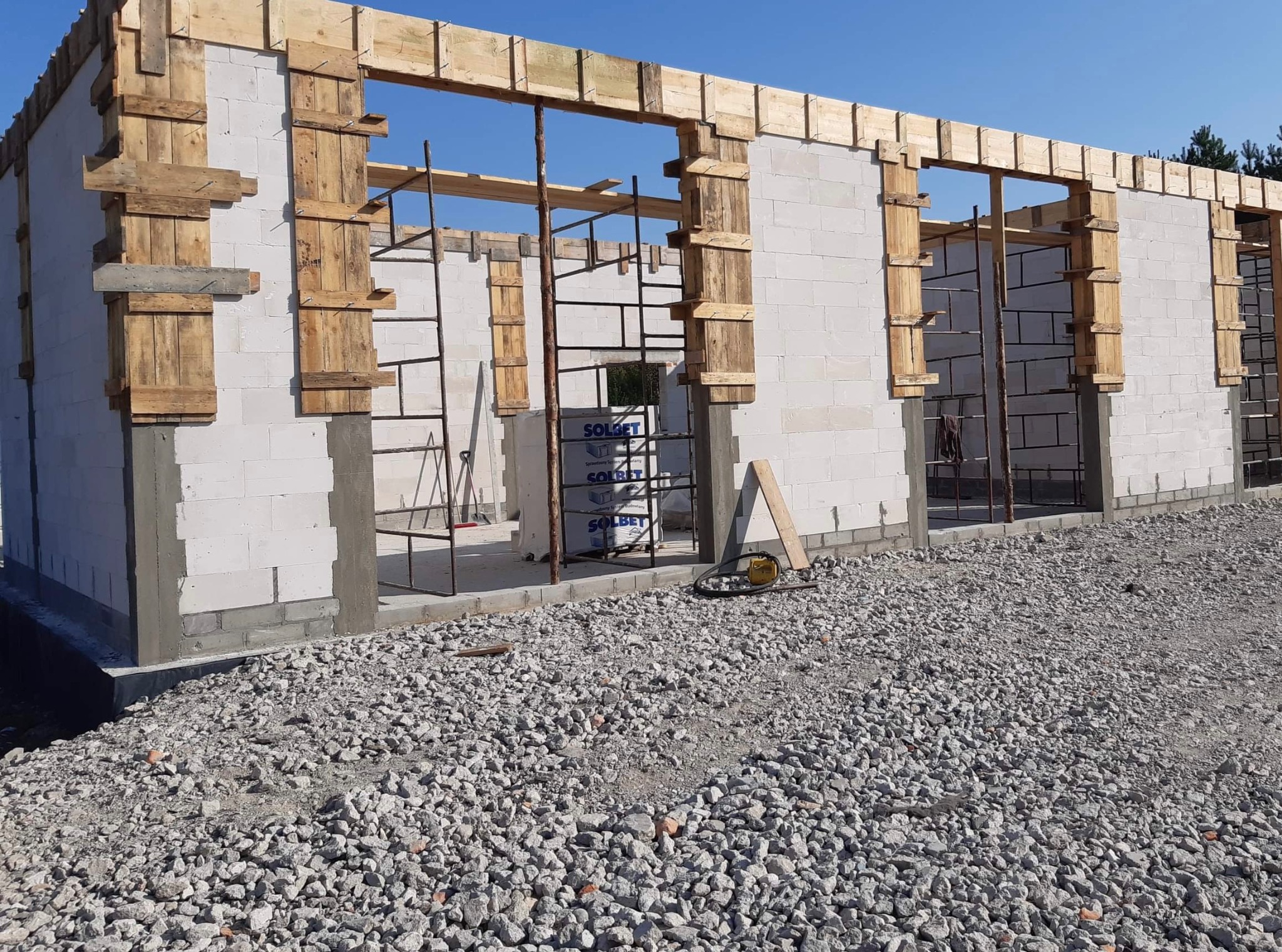 Budowa domu z białych bloczków: ściany w trakcie wznoszenia, widoczne drewniane szalunki okienne, stalowe rusztowania i paleta bloczków Solbet w tle.