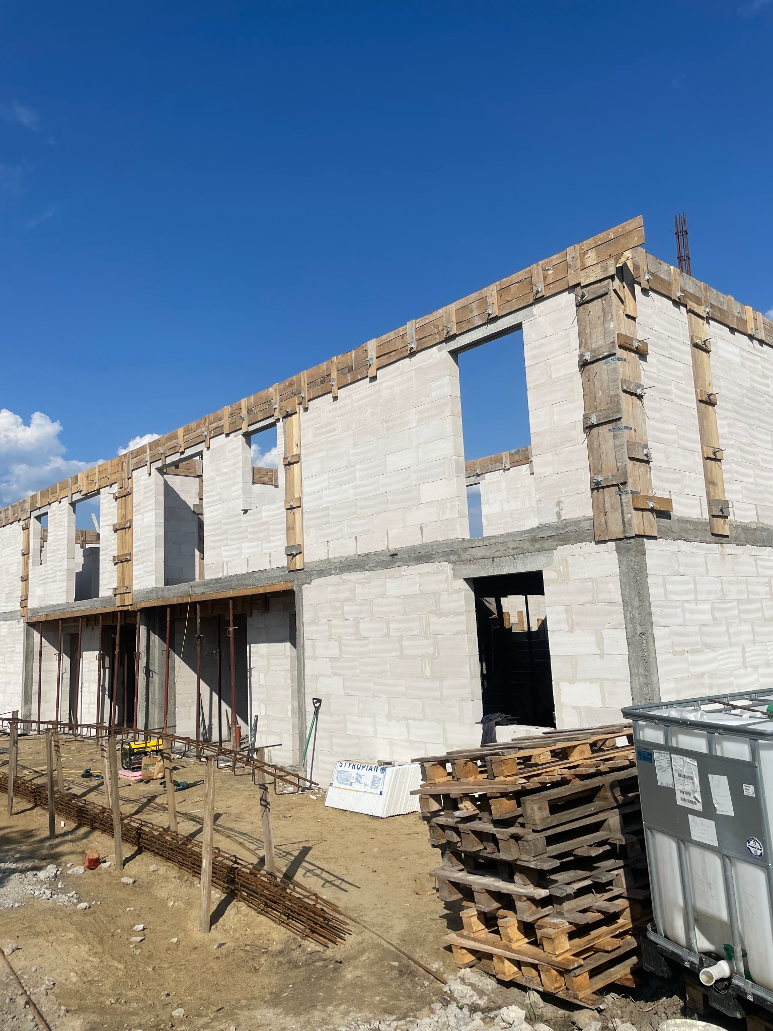 Surowy stan dwukondygnacyjnego domu z białych bloczków, z widocznymi drewnianymi szalunkami okiennymi i zbrojeniami, na tle błękitnego nieba, z paletami i zbiornikiem IBC u podstawy.
