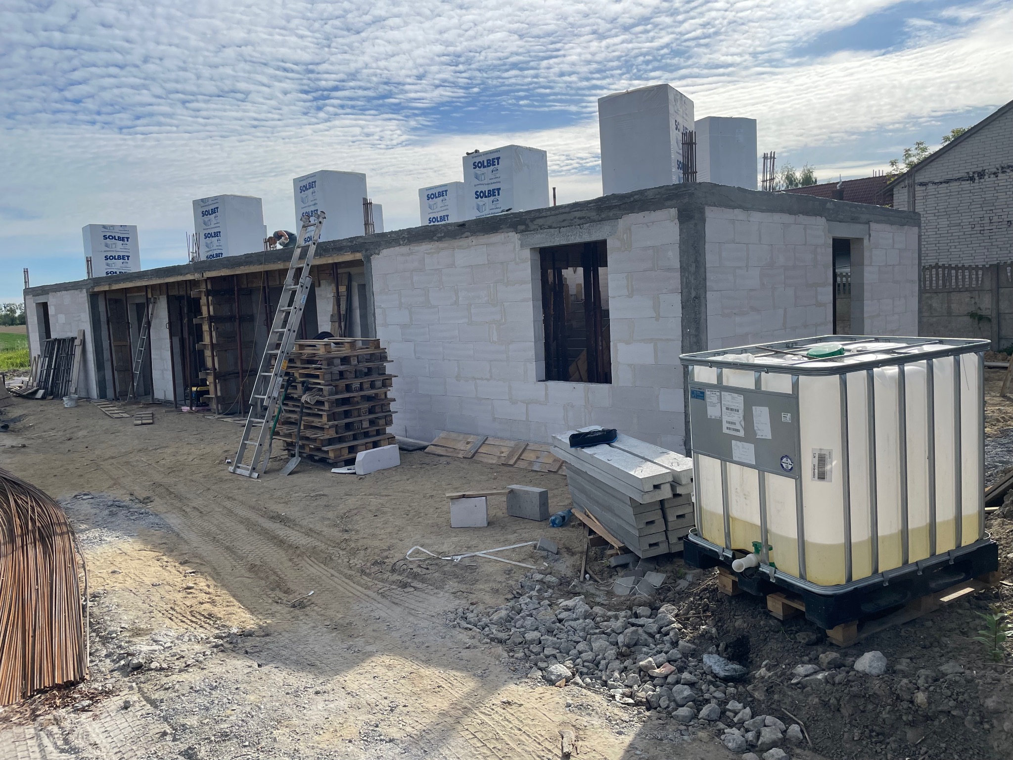 Wznoszenie ścian parterowego domu z bloczków Solbet, widoczne szalunki okienne, palety z materiałami i zbiornik na wodę na placu budowy.