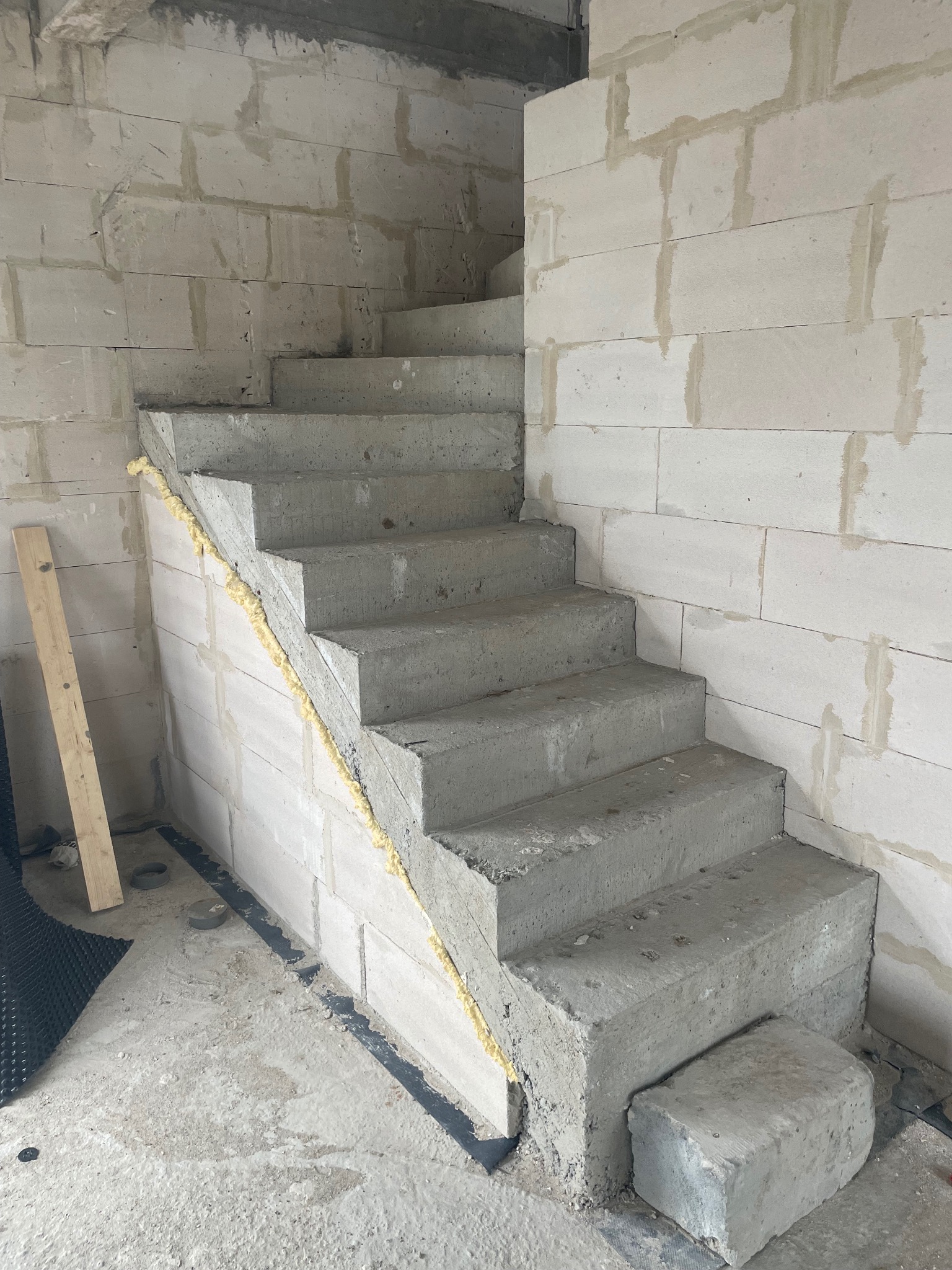 Surowe, betonowe schody wewnętrzne w trakcie budowy, widoczne bloki z betonu komórkowego, częściowo wypełniona pianką montażową szczelina między schodami a ścianą, oraz fragment folii fundamentowej...