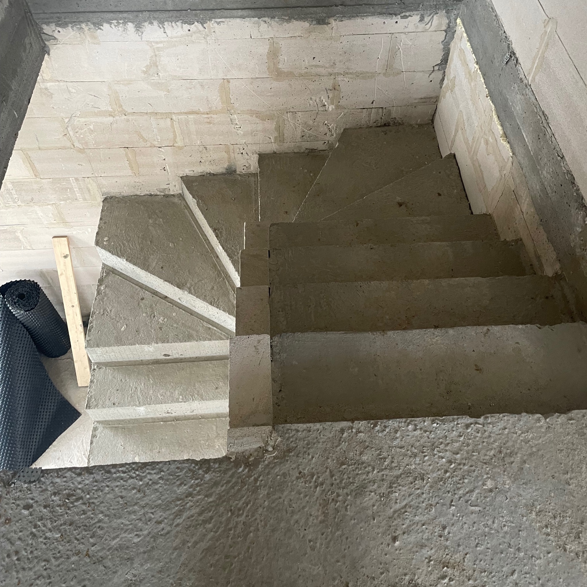 Wewnętrzne schody betonowe w surowym stanie, widok z góry, z rolką izolacji fundamentów i deską obok.