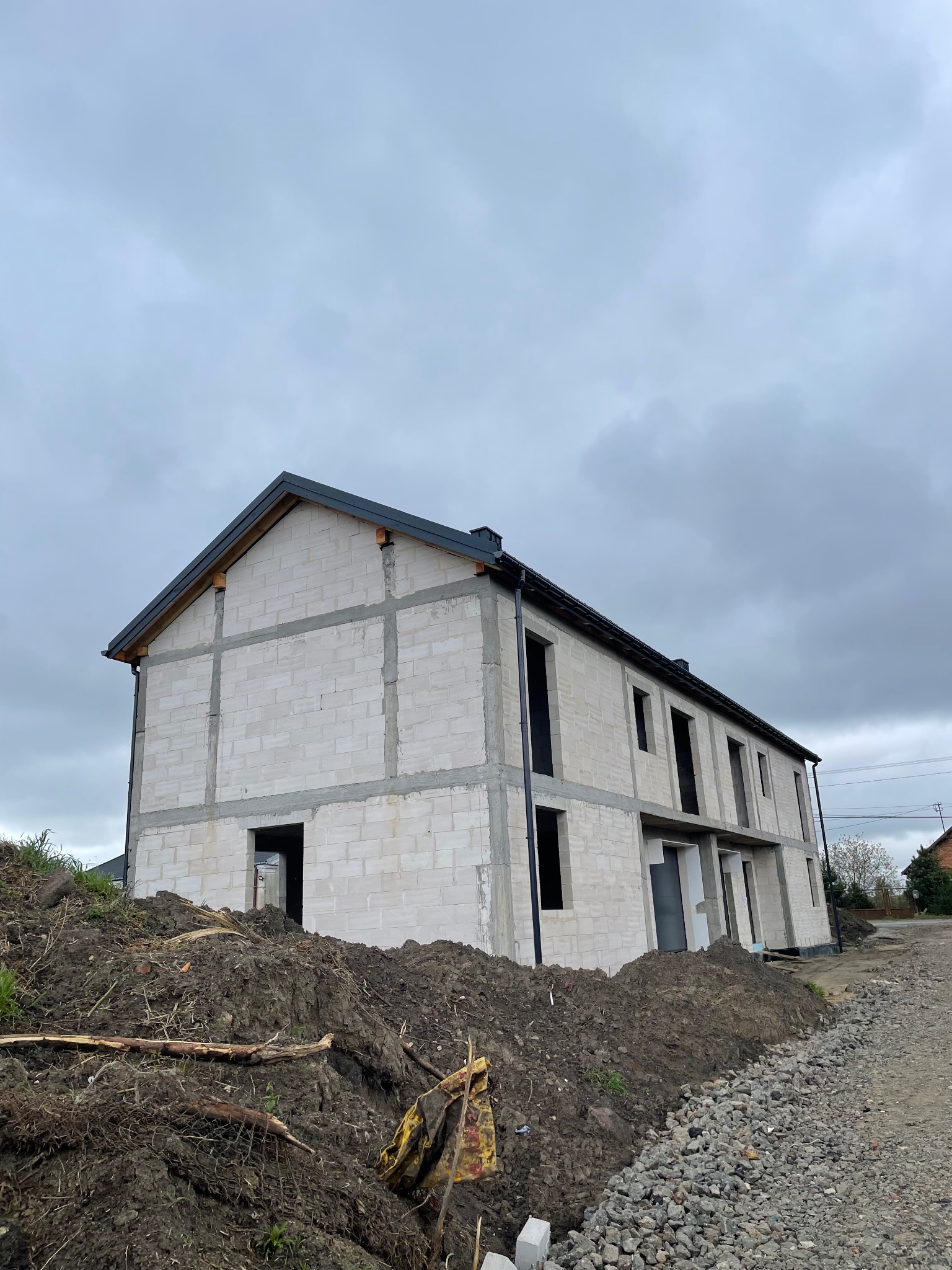 Dwukondygnacyjny dom w stanie surowym otwartym, zbudowany z białych bloczków, widoczne otwory okienne i drzwiowe, na pierwszym planie pryzma ziemi i gruzu, pochmurne niebo.