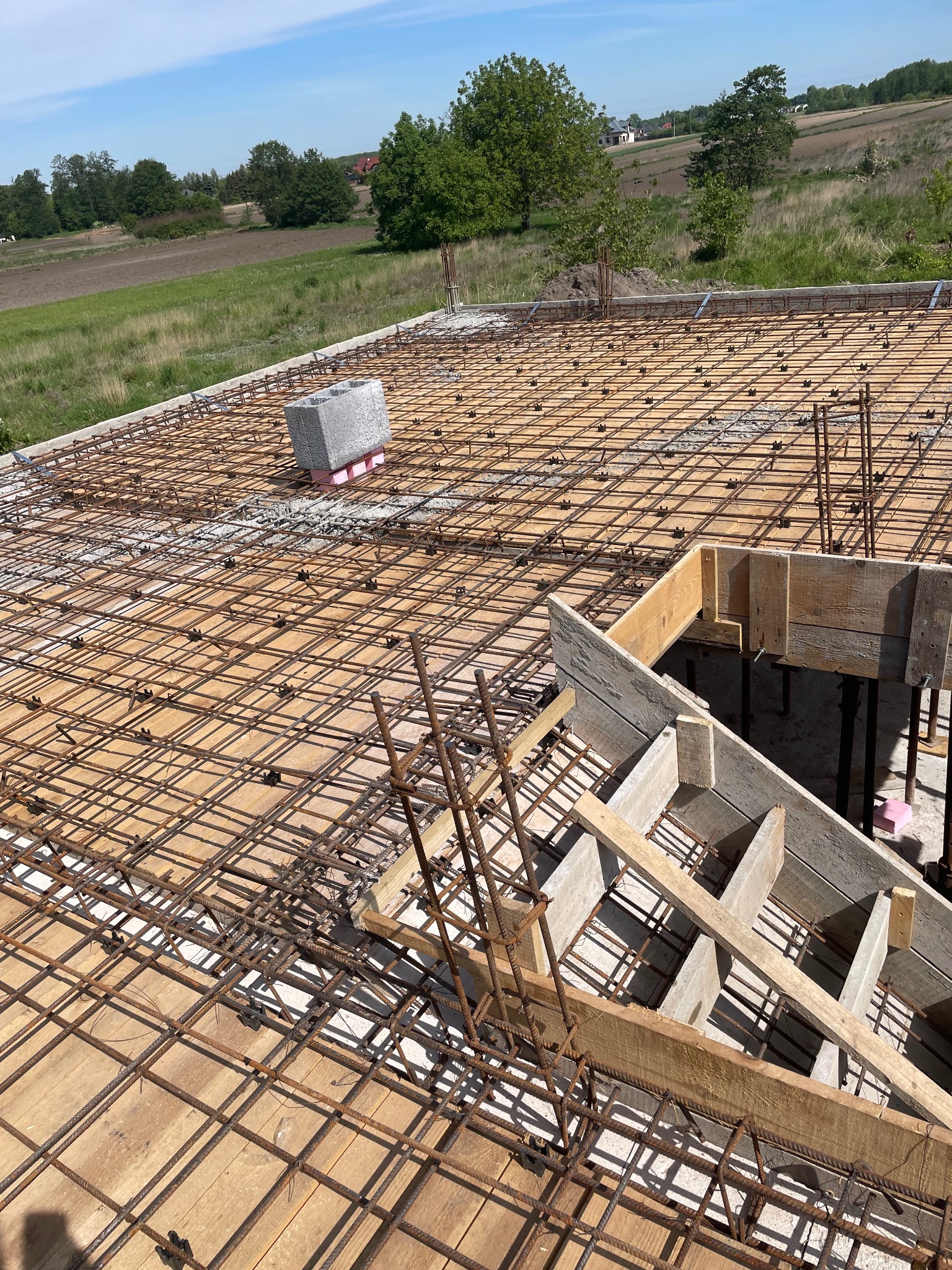 Zbrojenie stropu z drewnianym szalunkiem i widokiem na zielone pola w tle.