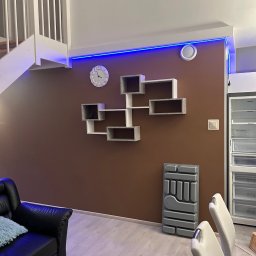 Tomasz Ejlak - Wnętrze pokoju z brązową ścianą, białymi półkami na ścianie, zegarem, czarną skórzaną kanapą z turkusową poduszką, białymi krzesłami i lodówką po prawej stronie, oświetlone niebieskimi lampkami LED.