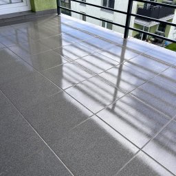 Tomasz Ejlak - Błyszcząca, szara podłoga z płytek na balkonie, z odbiciem metalowej balustrady i budynku w tle.