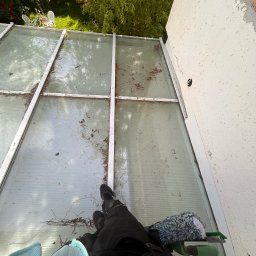 Tomasz Ejlak - Widok z góry na zabrudzoną, przeszkloną konstrukcję dachu z widocznymi liśćmi i gałązkami, obok ściana budynku oraz narzędzia i odzież osoby czyszczącej.