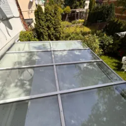 Przezroczysty dach z poliwęglanu z metalową ramą, widok na zielony ogród z drzewami, krzewami i pergolą w tle.