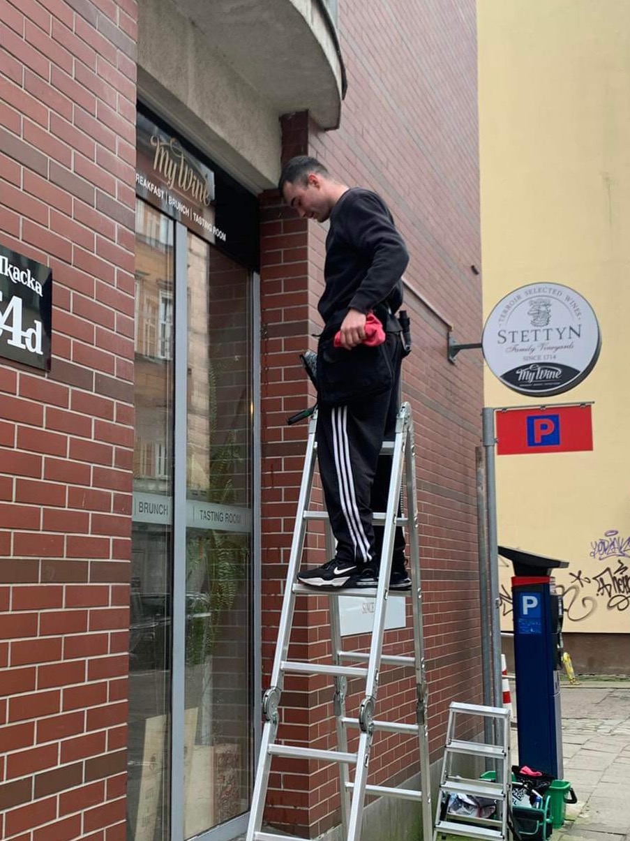 Pracownik w czarnym ubraniu na drabinie czyści szybę wejściową do lokalu 'My Wine', widoczny fragment ulicy z parkomatem i tablicą z adresem.