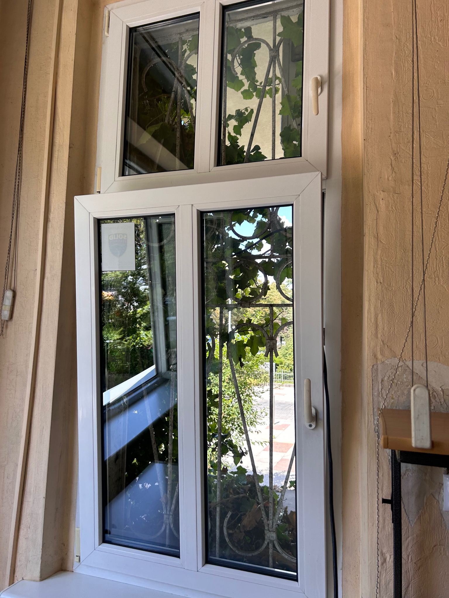 Otwarty biały plastikowy okno z widocznymi zielonymi liśćmi rośliny na zewnątrz i naklejką na szybie w górnym lewym rogu.