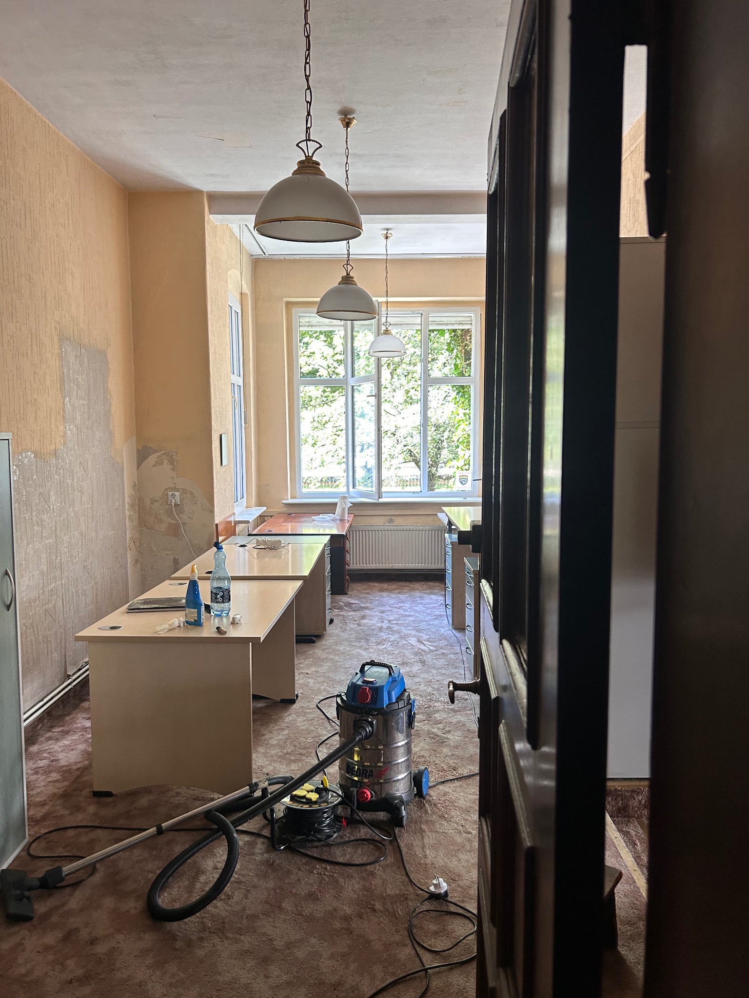 Wnętrze biura z widocznymi śladami niedawnego remontu, z odkurzaczem przemysłowym na pierwszym planie, butelką wody i środkiem czyszczącym na biurku, oraz fragmentem ściany z odsłoniętym tynkiem.