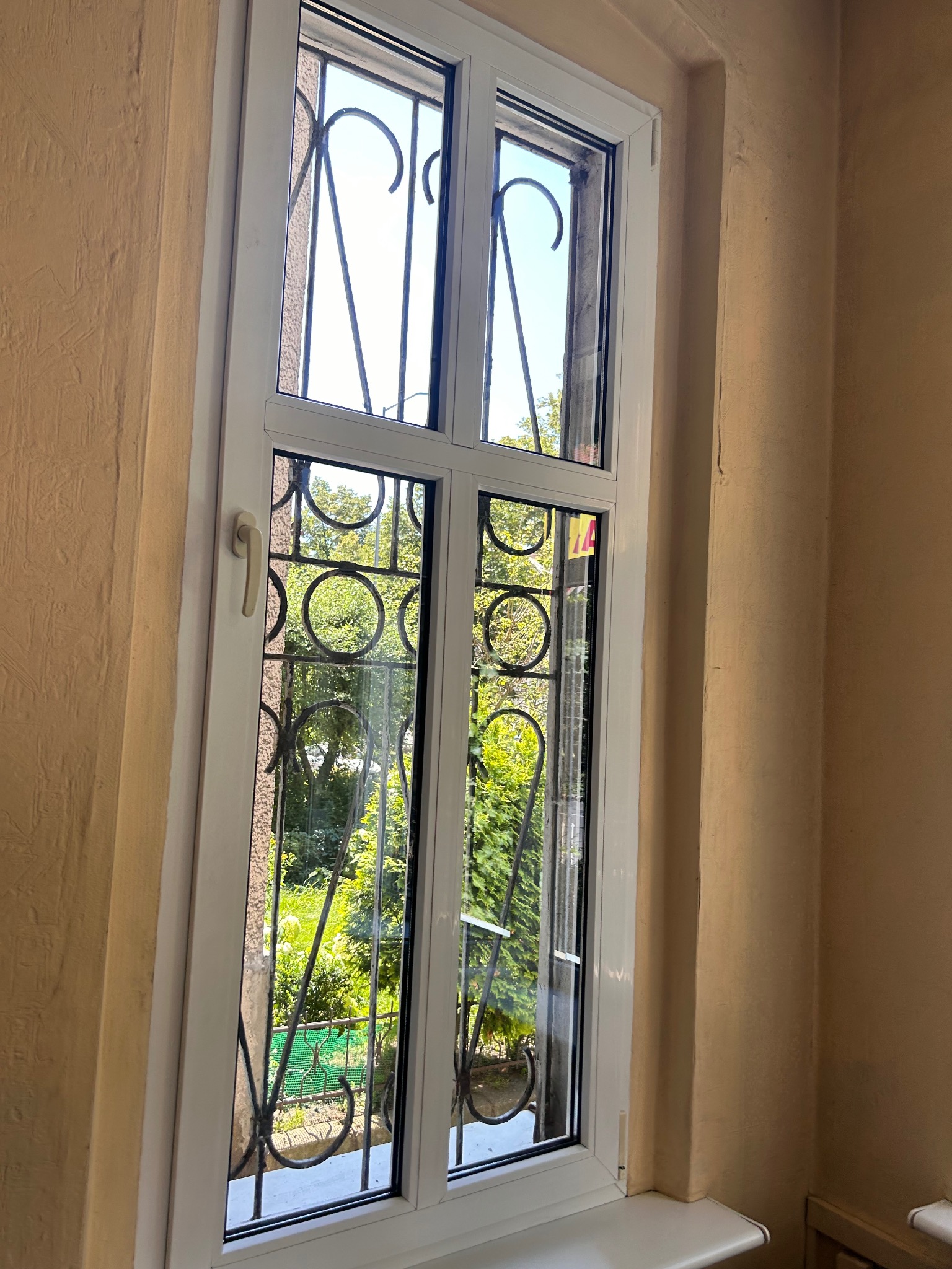 Białe okno PCV z ozdobną kratą wychodzące na zielony ogród, widoczne na tle beżowych ścian z fakturą.