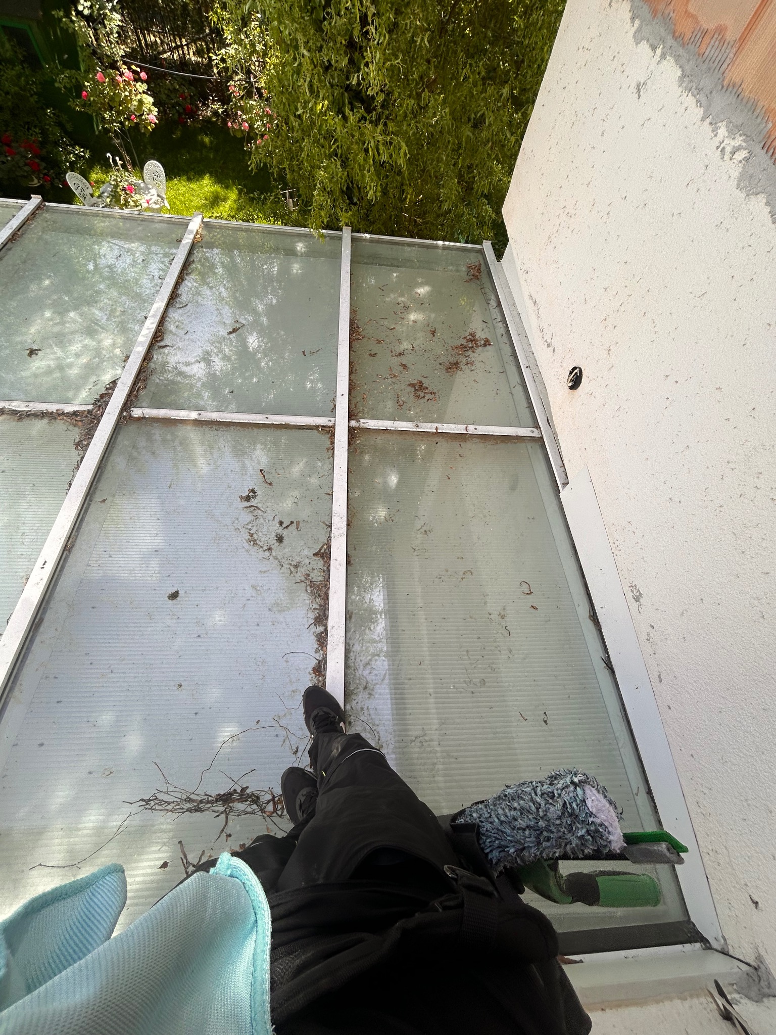 Widok z góry na zabrudzoną, przeszkloną konstrukcję dachu z widocznymi liśćmi i gałązkami, obok ściana budynku oraz narzędzia i odzież osoby czyszczącej.