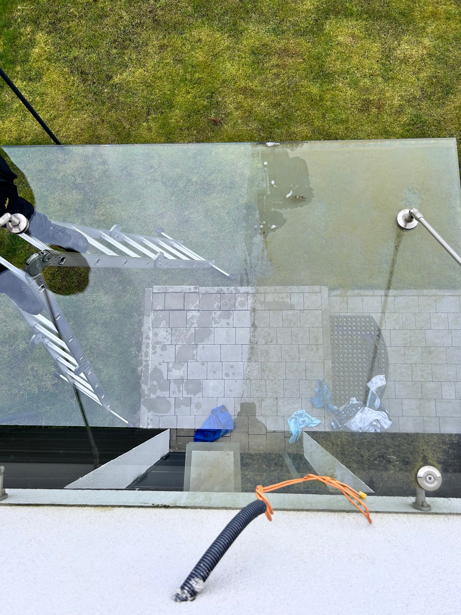 Widok z góry na szklany dach patio podczas czyszczenia, z widocznymi drabinami, brudną powierzchnią, narzędziami i elementami otoczenia, takimi jak trawa i kostka brukowa.