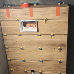 Ściana wyłożona płytkami imitującymi drewno, z widocznymi elementami instalacji elektrycznej i hydraulicznej, zabezpieczona pomarańczową taśmą i klipsami do płytek podczas montażu.