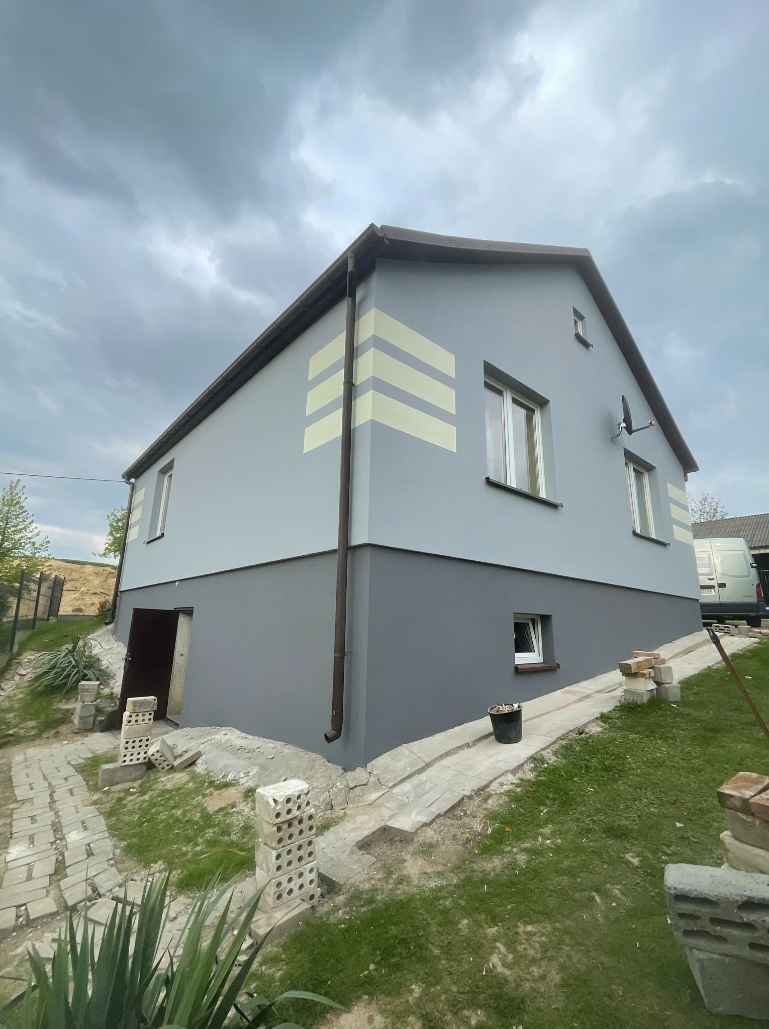 Dwukondygnacyjny dom z szarą elewacją, pomalowany w dwa odcienie szarości z poziomymi, jasnymi pasami dekoracyjnymi, widoczny z zewnątrz, z elementami krajobrazu i budowy.