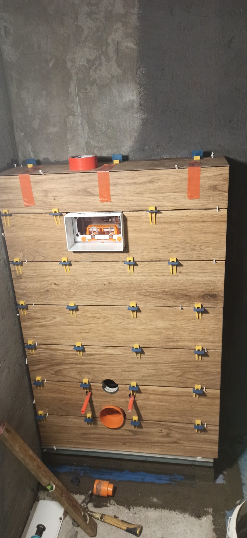 Ściana wyłożona płytkami imitującymi drewno, z widocznymi elementami instalacji elektrycznej i hydraulicznej, zabezpieczona pomarańczową taśmą i klipsami do płytek podczas montażu.
