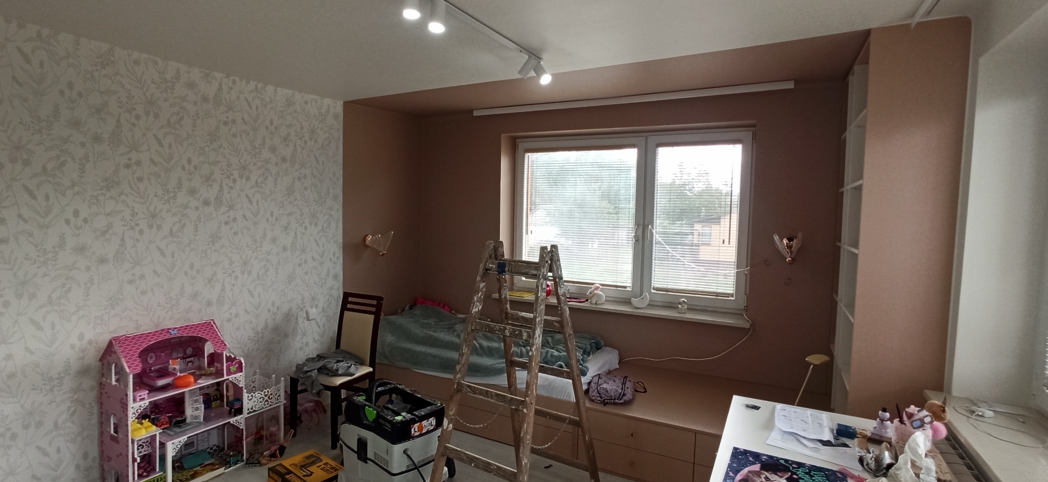 Pokój dziecięcy w trakcie remontu: ściana z tapetą w kwiaty, pomalowana na brązowo wnęka z oknem, drabina malarska oparta o wnękę, zabawki i narzędzia remontowe.