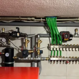 Rozdzielacz ogrzewania podłogowego z zielonymi rurkami, widoczne również pompy obiegowe i manometry, instalacja zamontowana na ścianie z płytek ceramicznych.