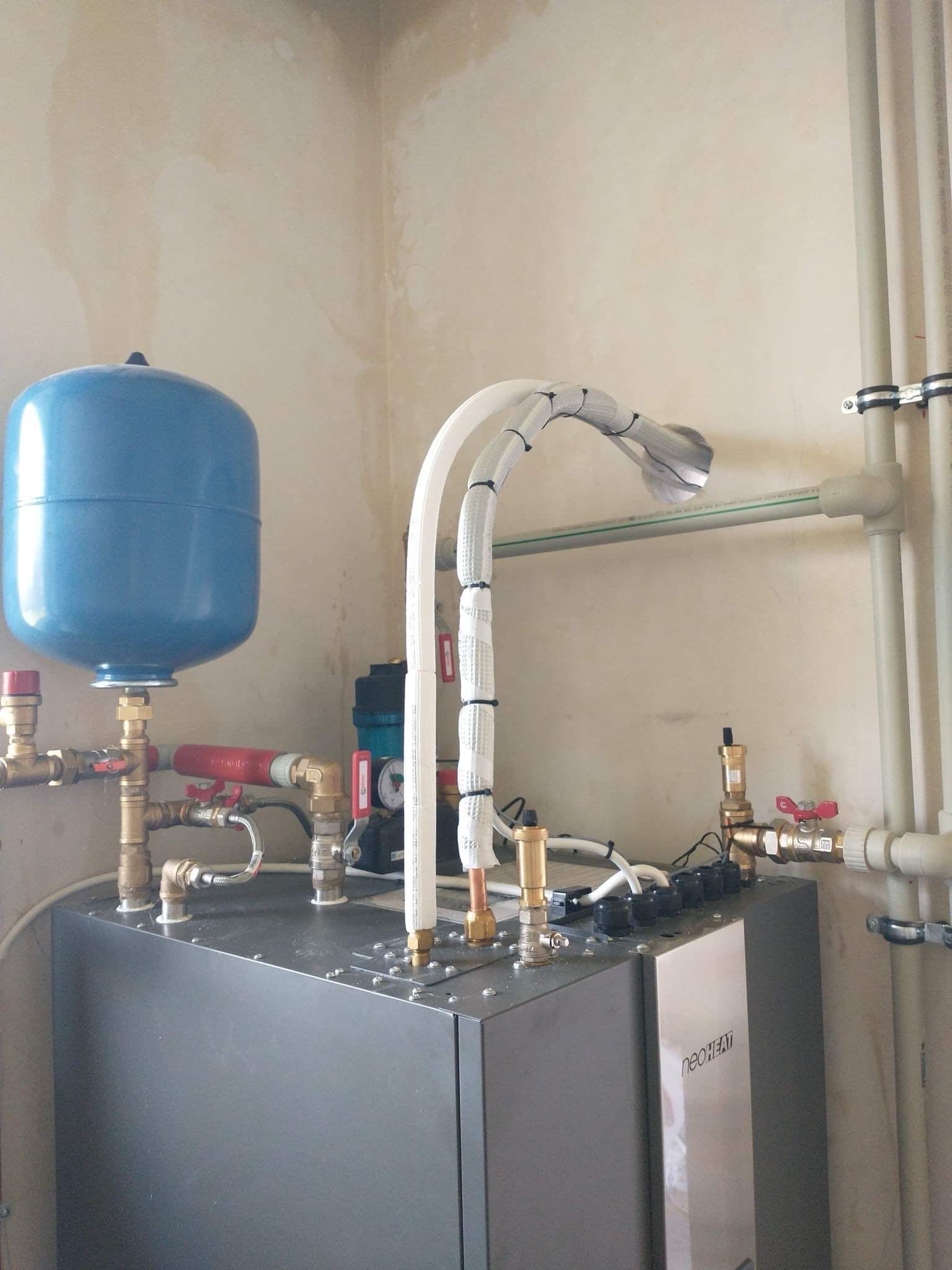 Szary piec grzewczy NeoHeat z niebieskim zbiornikiem wyrównawczym i rurami w białej izolacji, zamontowany w pomieszczeniu z niedokończonymi ścianami.