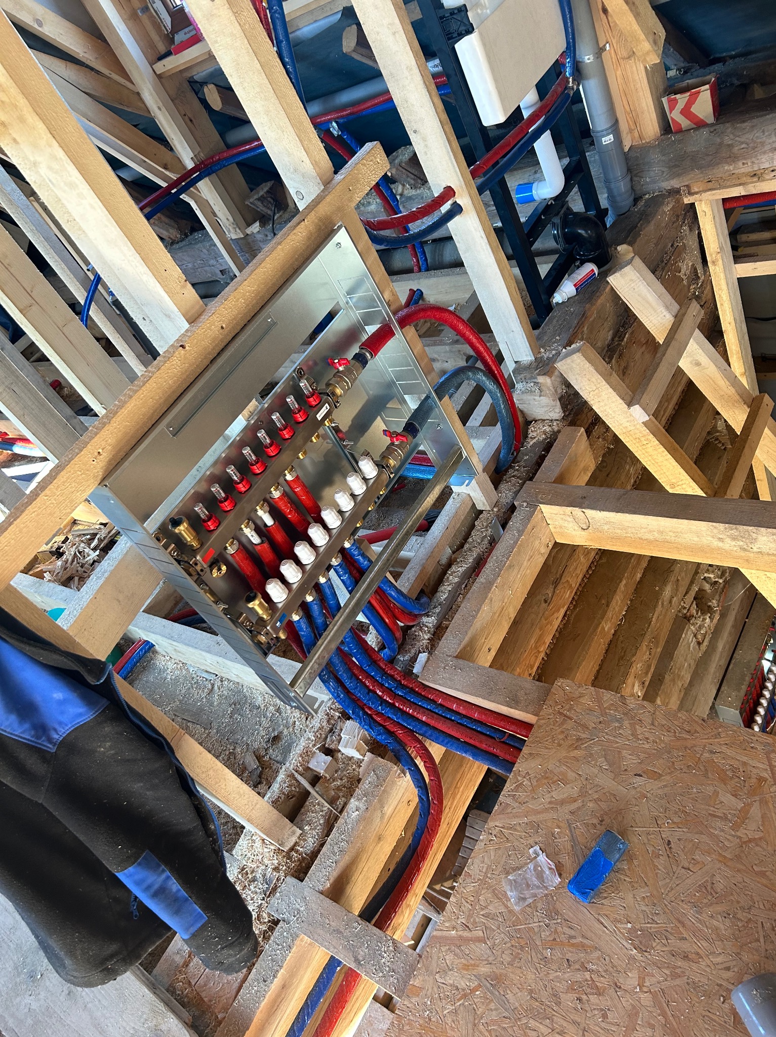 Rozdzielacz ogrzewania podłogowego zamontowany w drewnianej konstrukcji stropu, widoczne rury w kolorze czerwonym i niebieskim oraz elementy instalacji wodnej i kanalizacyjnej.
