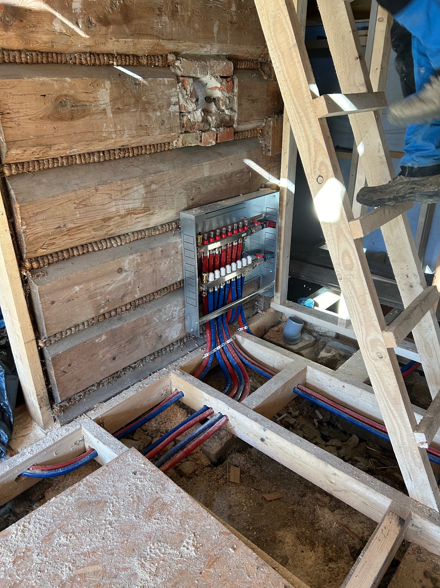 Rozdzielacz ogrzewania podłogowego zamontowany na ścianie z bali, z widocznymi rurami rozprowadzonymi w drewnianej konstrukcji podłogi, drabina oparta o ścianę.