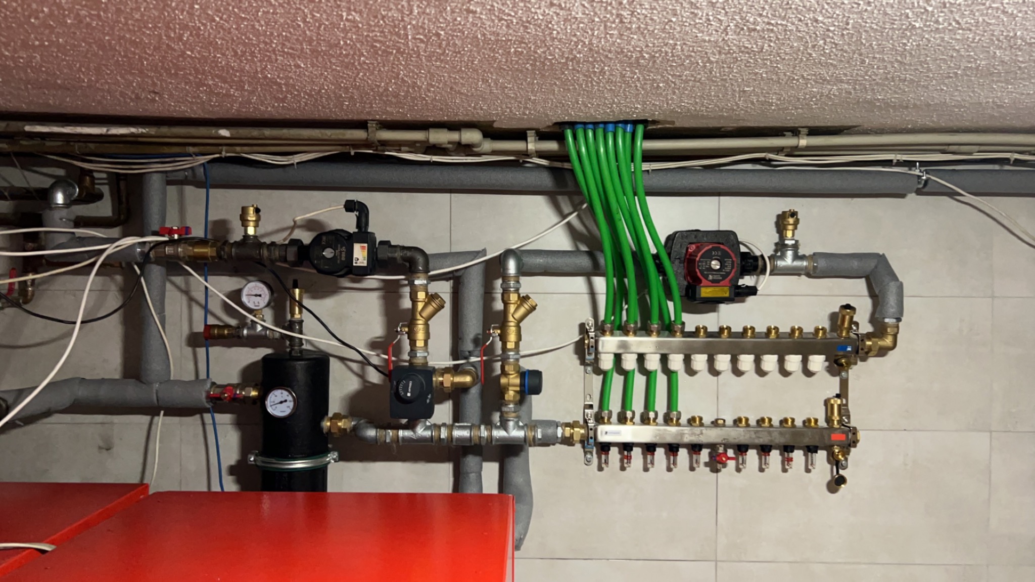Rozdzielacz ogrzewania podłogowego z zielonymi rurkami, widoczne również pompy obiegowe i manometry, instalacja zamontowana na ścianie z płytek ceramicznych.
