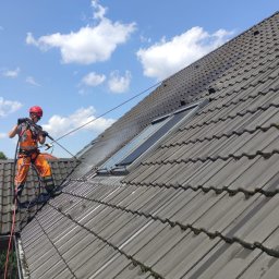 Mycie Ciśnieniowe Marcel Libera - Pracownik w pomarańczowym kombinezonie, zabezpieczony linami, czyści dach z szarej dachówki za pomocą myjki ciśnieniowej; widoczne okna dachowe i fragmenty mchu na dachówkach.