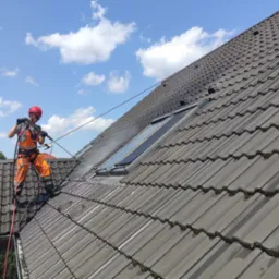 Pracownik w pomarańczowym kombinezonie, zabezpieczony linami, czyści dach z szarej dachówki za pomocą myjki ciśnieniowej; widoczne okna dachowe i fragmenty mchu na dachówkach.