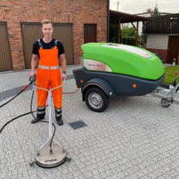 Mycie Ciśnieniowe Marcel Libera - Pracownik w pomarańczowym kombinezonie prezentuje urządzenie do czyszczenia kostki brukowej na tle domu z czerwonej cegły i zielonego urządzenia ciśnieniowego na przyczepie.
