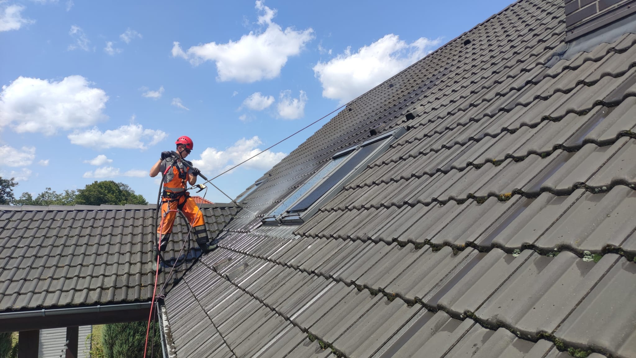 Pracownik w pomarańczowym kombinezonie, zabezpieczony linami, czyści dach z szarej dachówki za pomocą myjki ciśnieniowej; widoczne okna dachowe i fragmenty mchu na dachówkach.