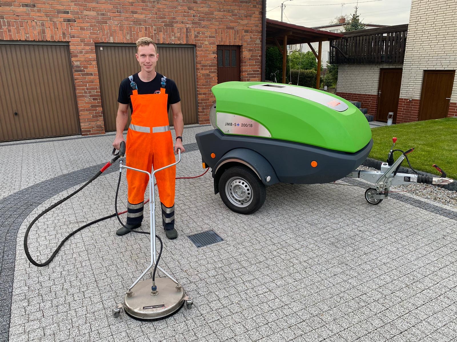 Pracownik w pomarańczowym kombinezonie prezentuje urządzenie do czyszczenia kostki brukowej na tle domu z czerwonej cegły i zielonego urządzenia ciśnieniowego na przyczepie.