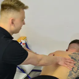 Mężczyzna w czarnej koszulce wykonuje masaż pleców wytatuowanemu mężczyźnie leżącemu na leżance w gabinecie.